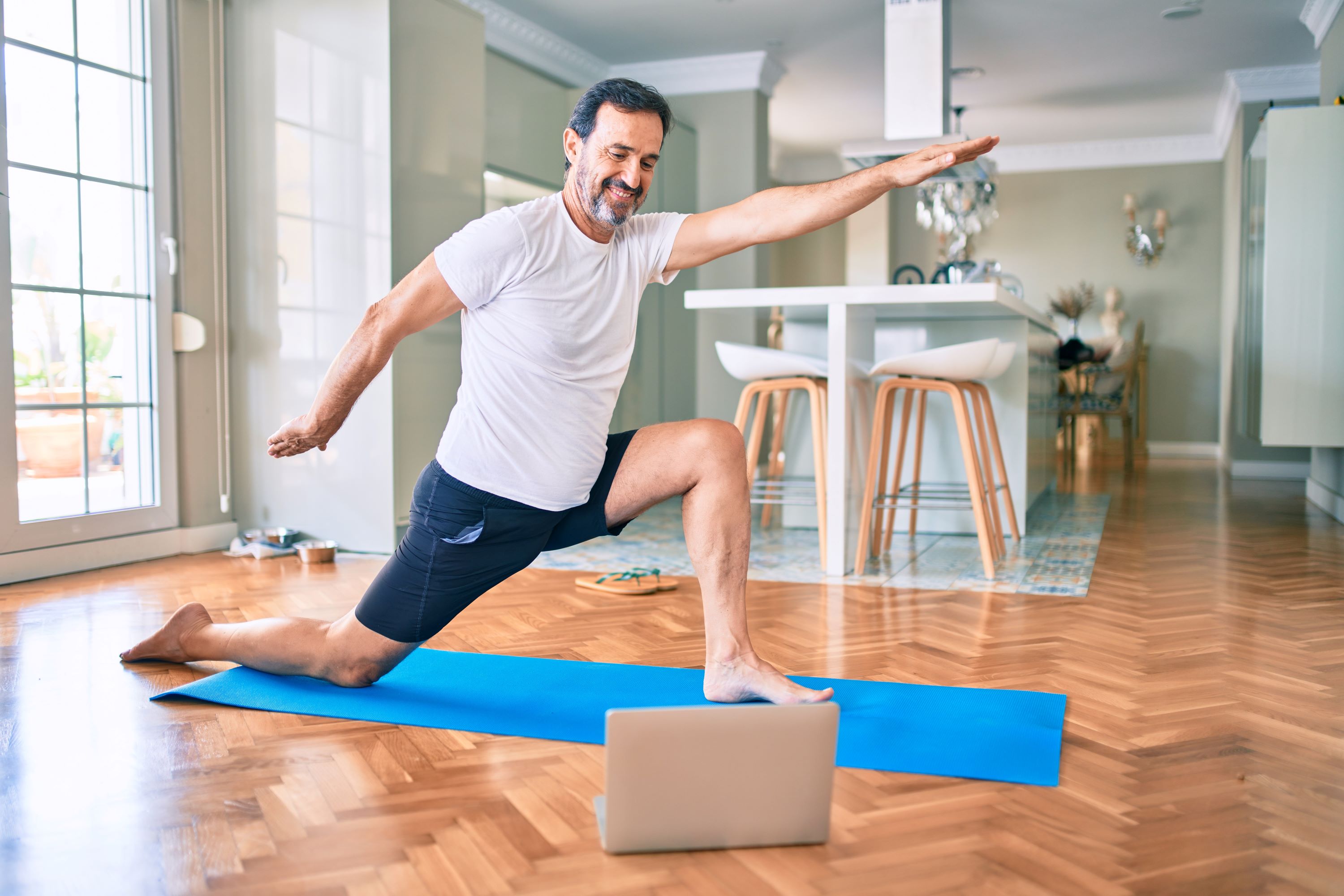 man doing yoga at home wearing white shirt on blue yoga mat and wooden floor hand raised in air