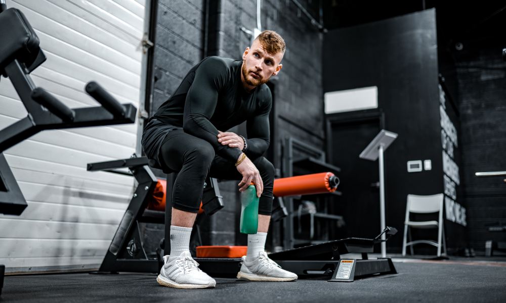 guy sitting on bench at gym holding water flask wearing black with white sneakers