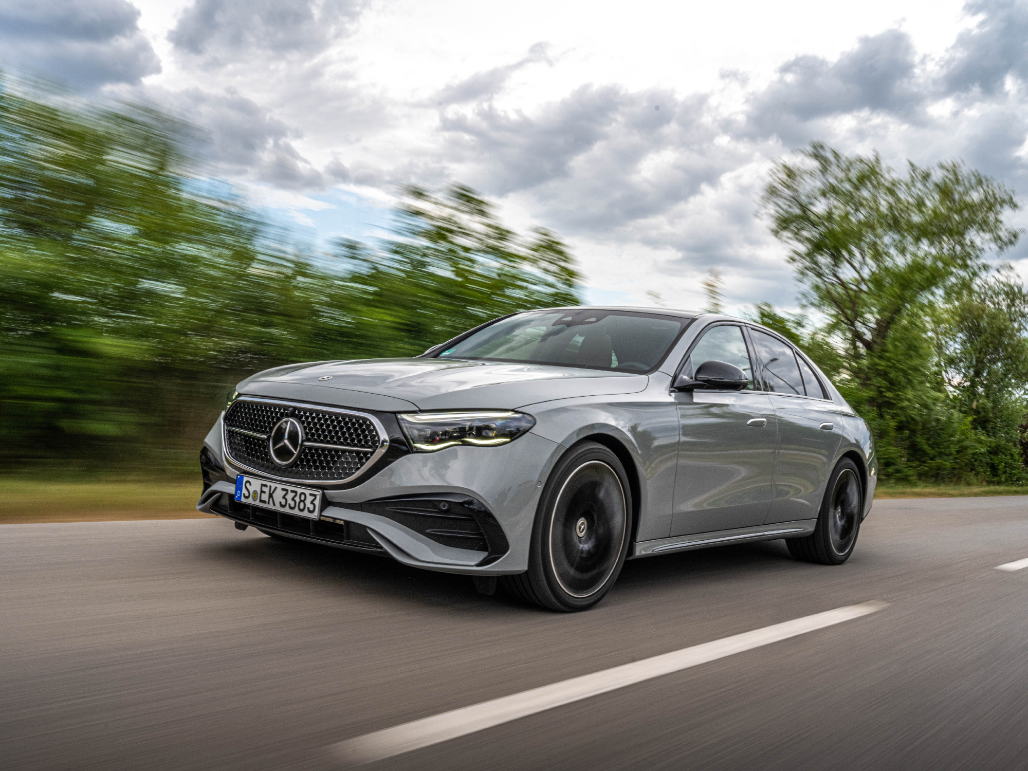 2024 Mercedes-Benz E-Class Sedan left front three-quarter shot driving on road with tress and a cloudy sky in the background.