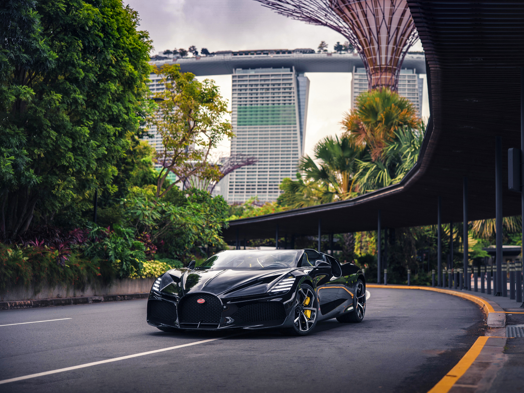 A black Bugatti W16 Mistral Roadster driving on a street in Singapore with heavy vegetation beside the road and modernistic structures overhead.