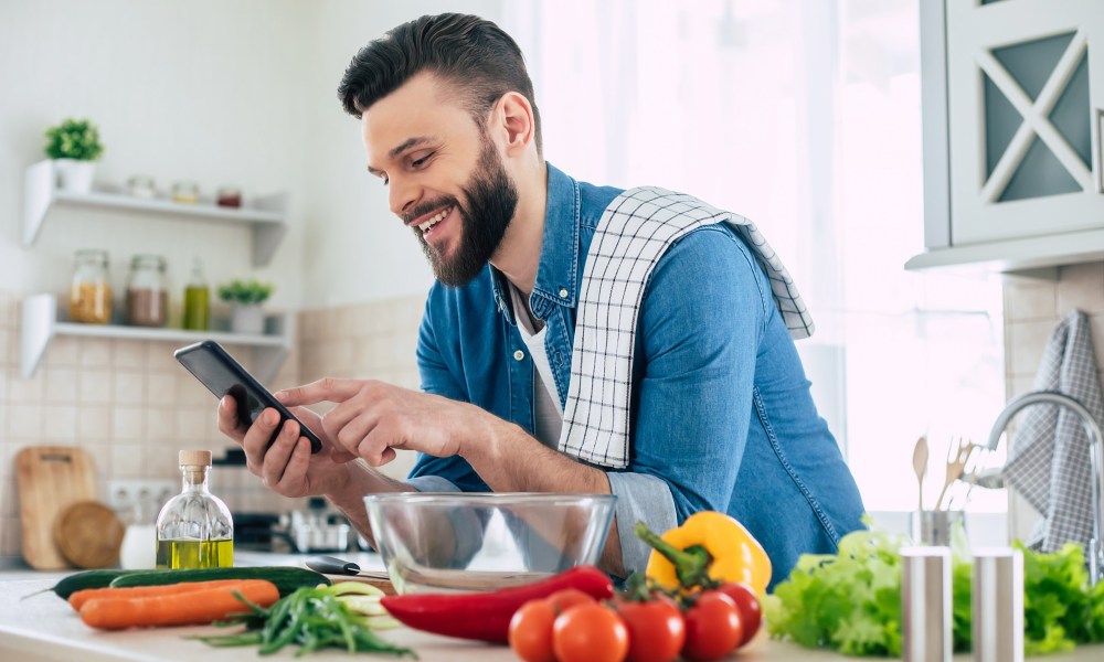 Happy handsome bearded man is using his smart phone while he preparing vegan healthy breakfast for a lovely family