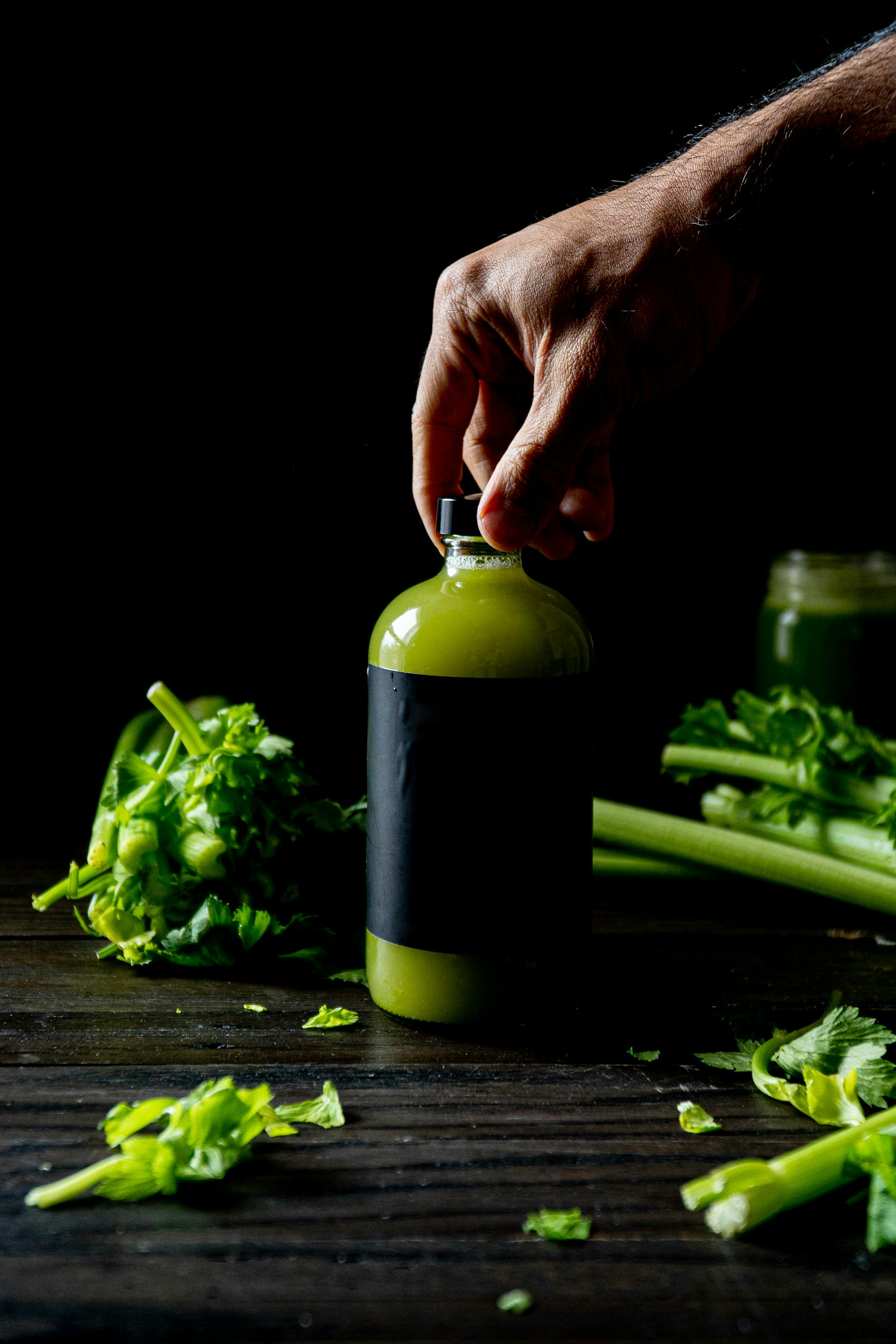 someone's hand holding the bottle of green drink celery juice on dark background with green celery stalks