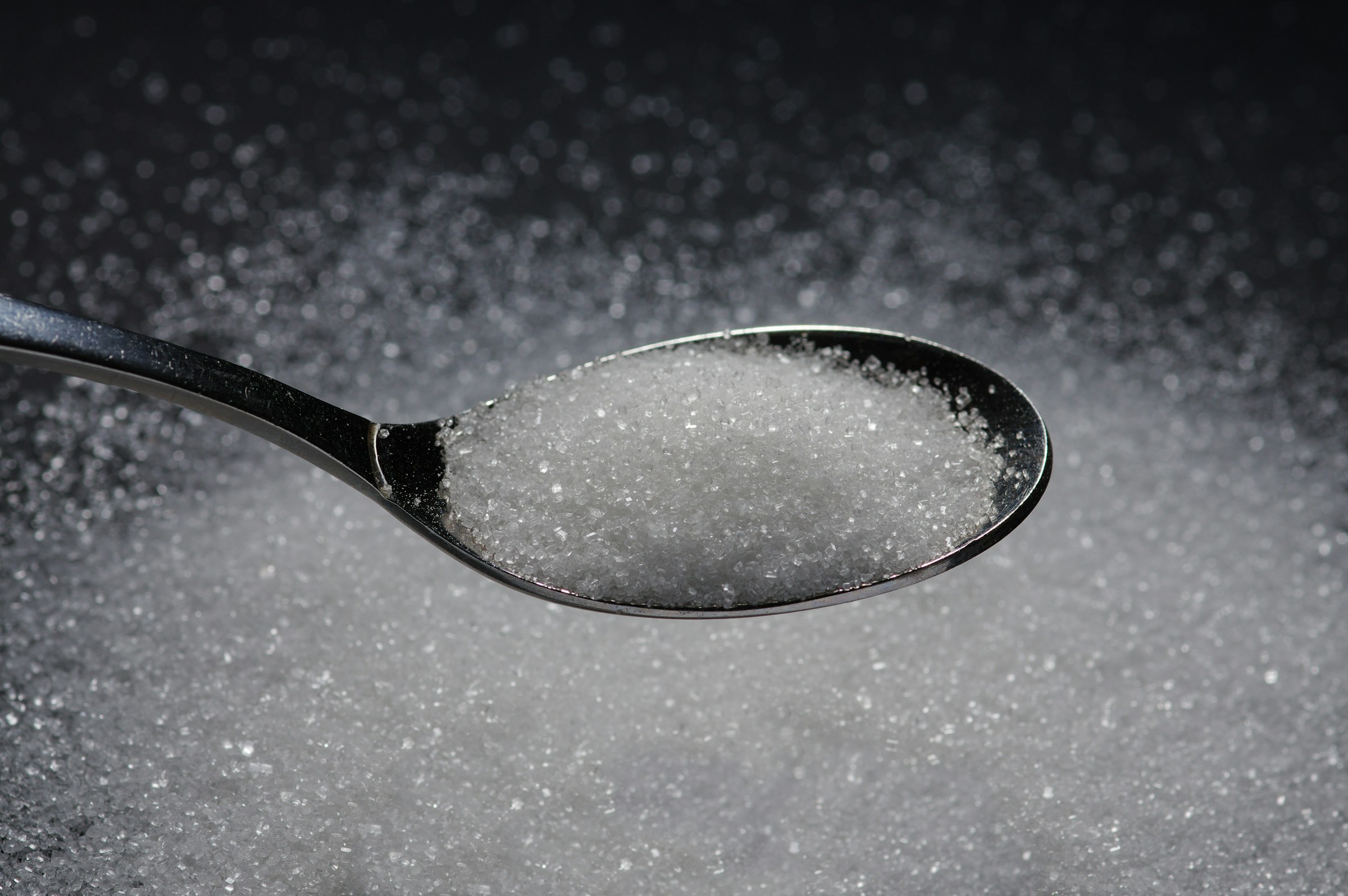 spoonful of white sugar held above a dark table with white sugar on the table