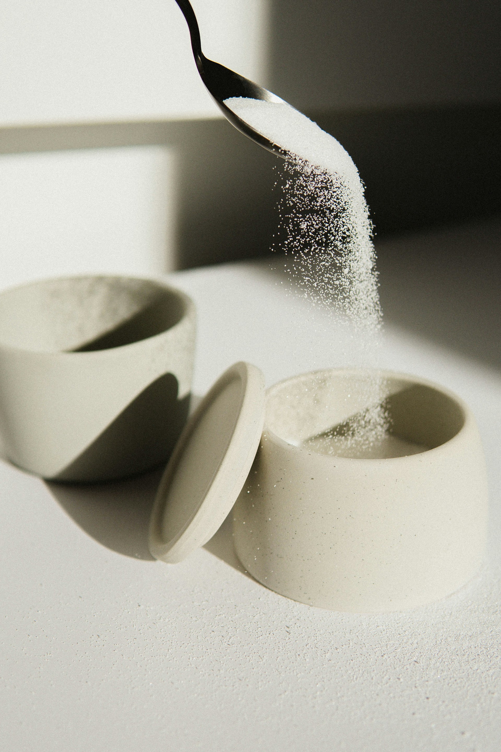 pouring white sugar from silver metallic spoon into white ceramic bowl on table