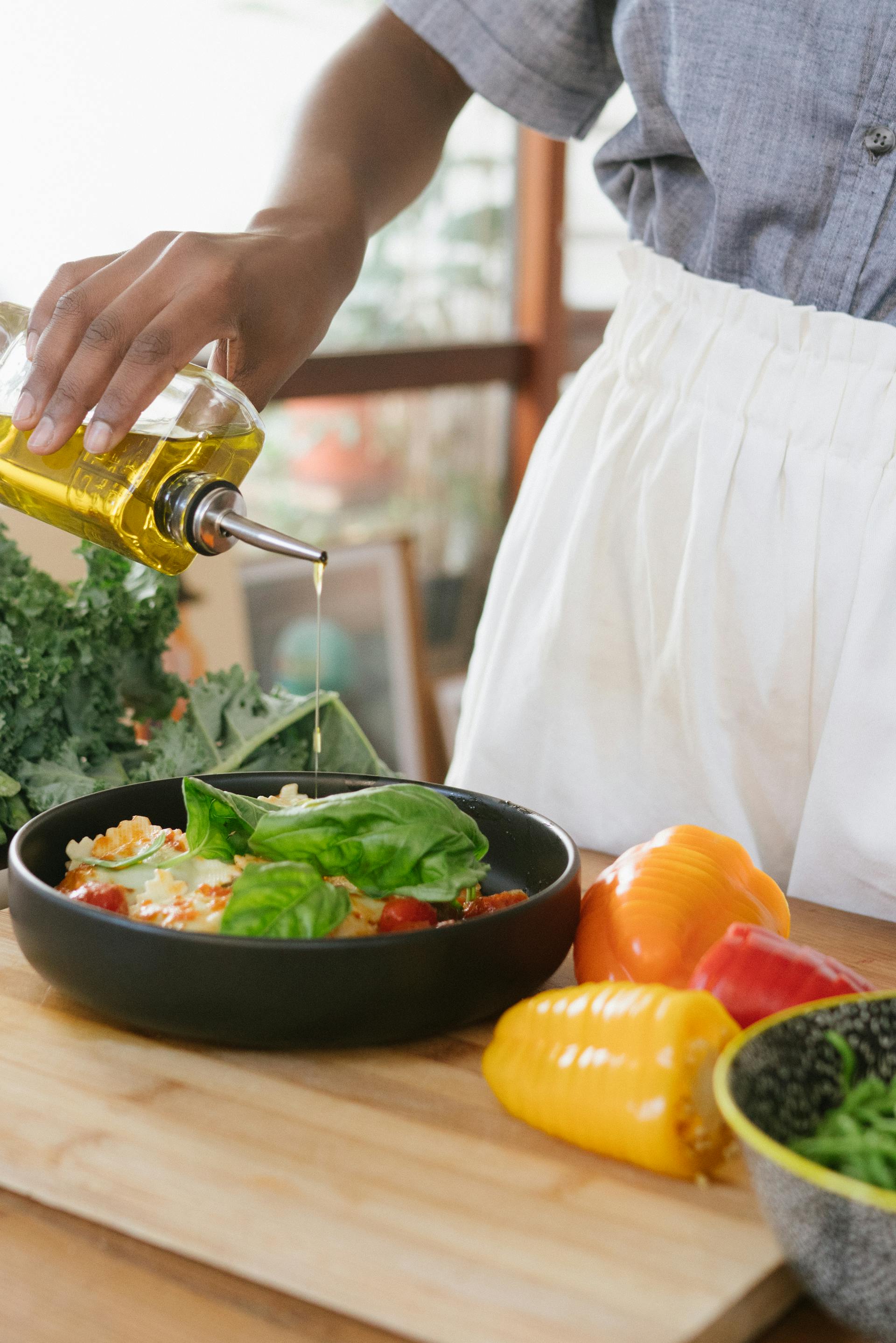 Cooking pouring olive oil onto black pan with veggies and herbs on wooden table