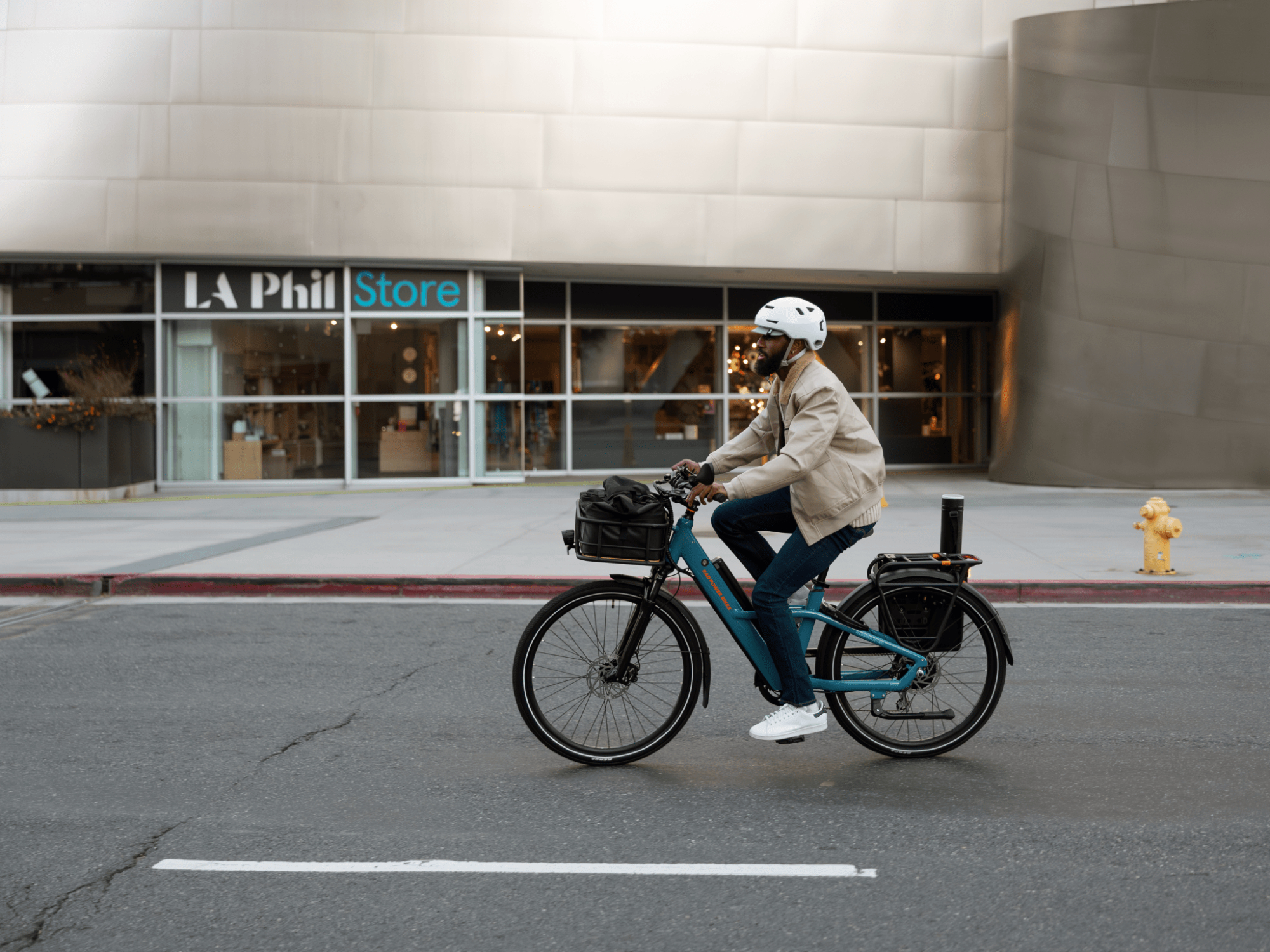 A person riding a Rad Power Bikes Radster Road electric commuter e-bike on a city street passing in front of a store entrance.