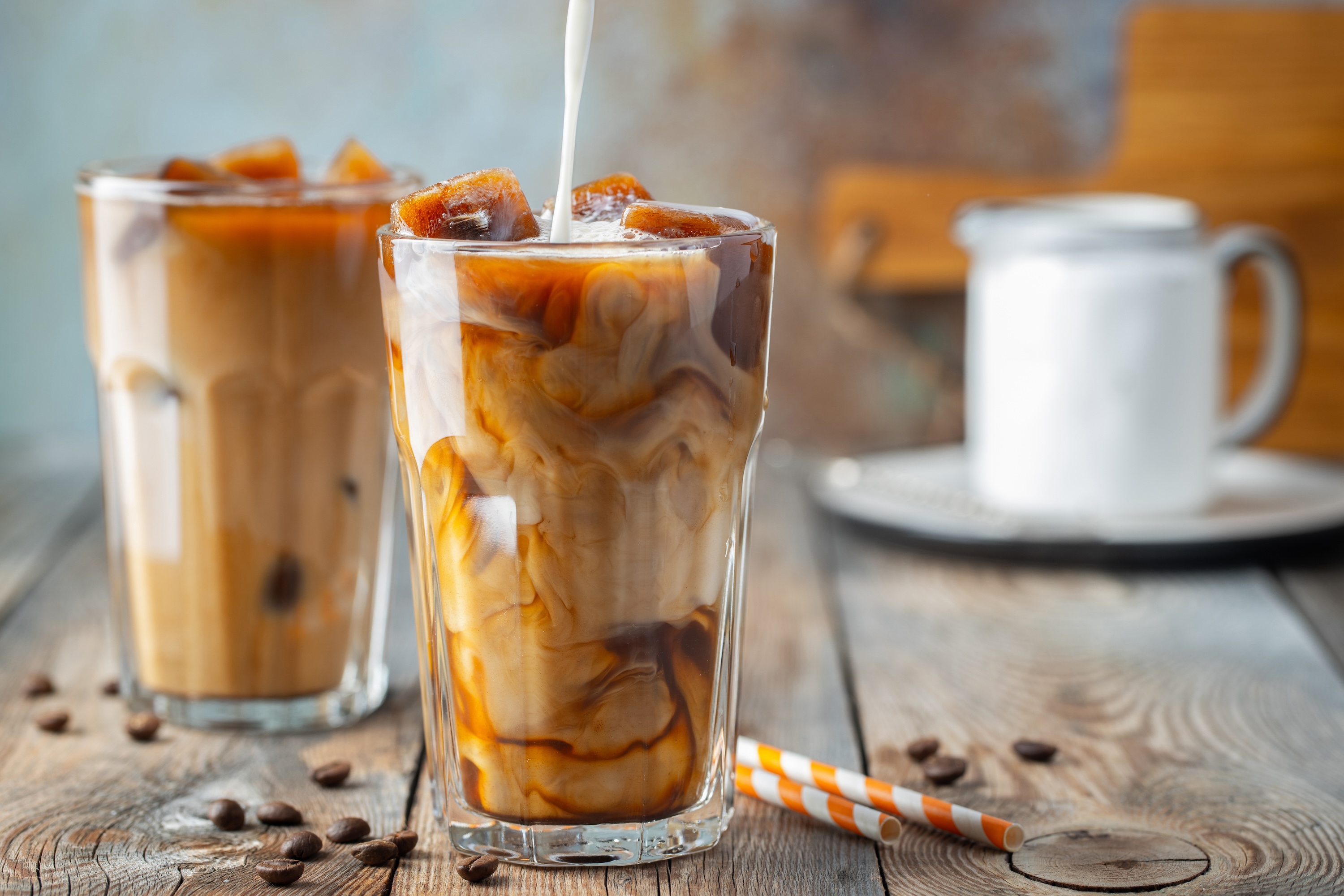 Ice coffee in a tall glass with cream poured over, coffee ice cubes and beans on a old rustic wooden table. Cold summer drink with tubes on a blue rusty background with copy space.