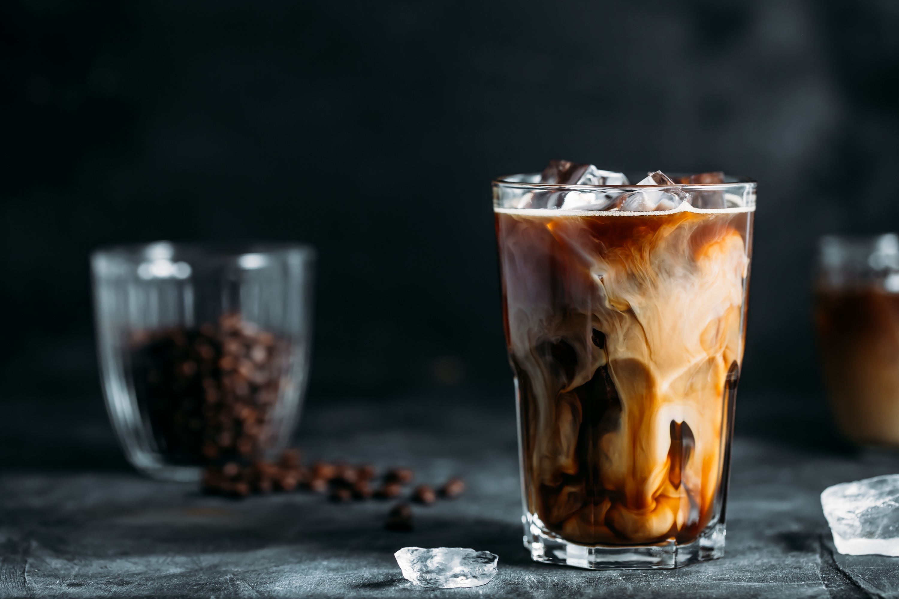 Ice coffee in a tall glass with cream poured over and coffee beans on dark gray background