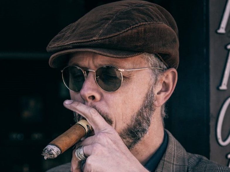 Man smoking a cigar in a newsboy cap outside.