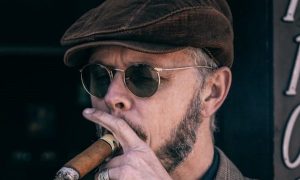 Man smoking a cigar in a newsboy cap outside.