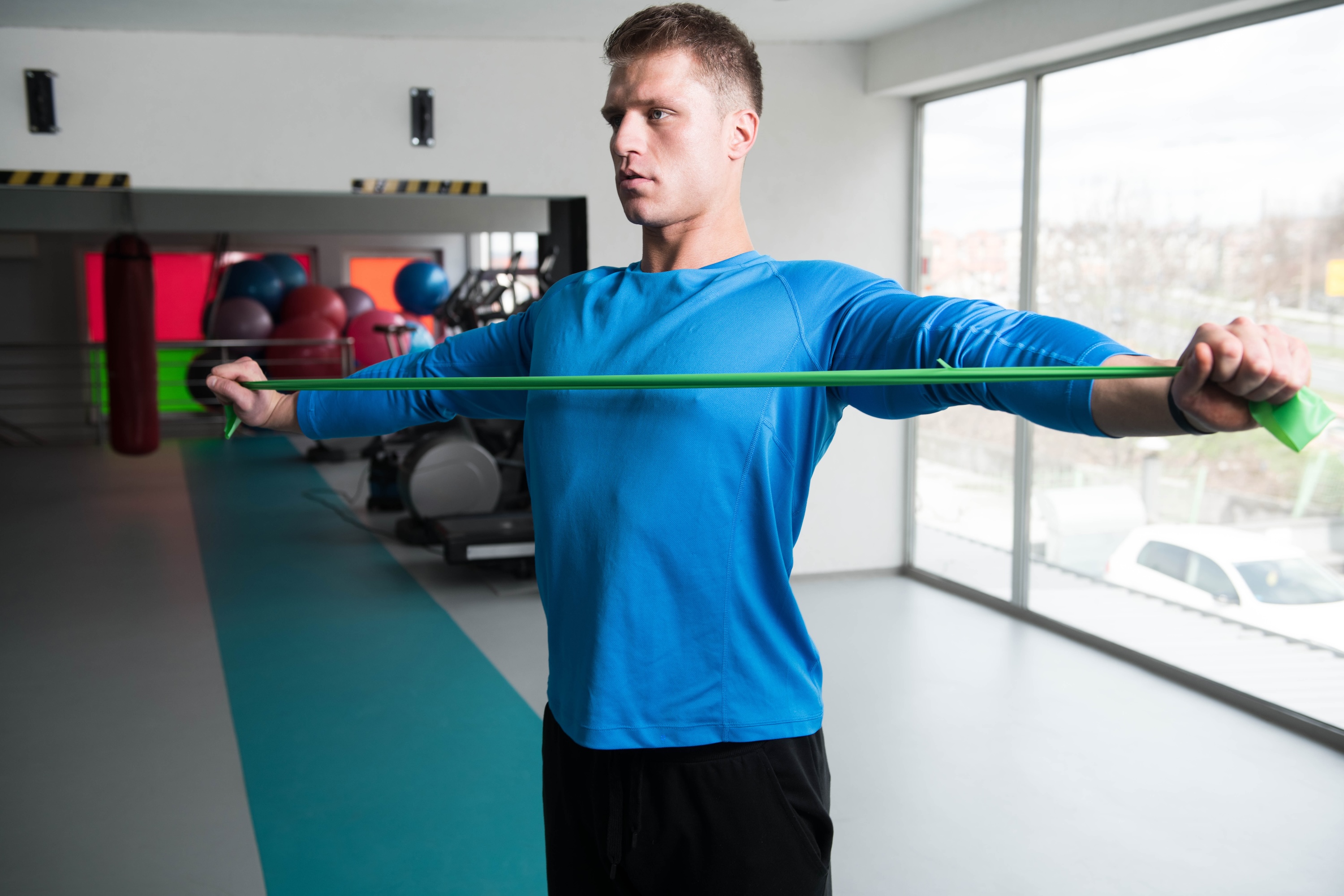 man wearing blue shirt stretching with resistance band performing band pull apart exercise in gym