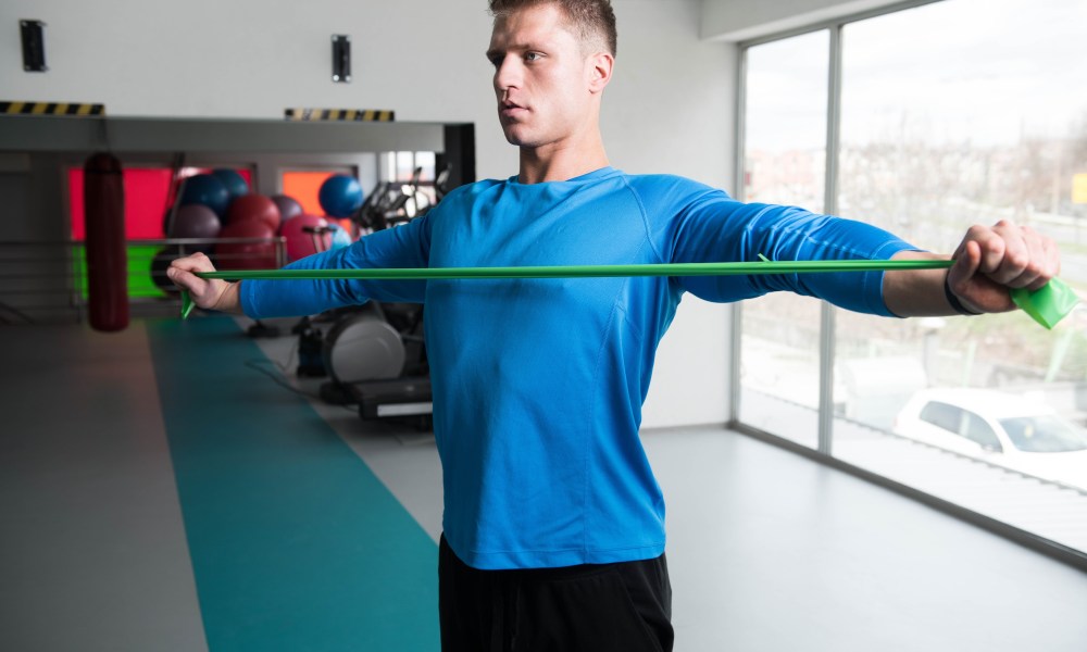 man wearing blue shirt stretching with resistance band performing band pull apart exercise in gym