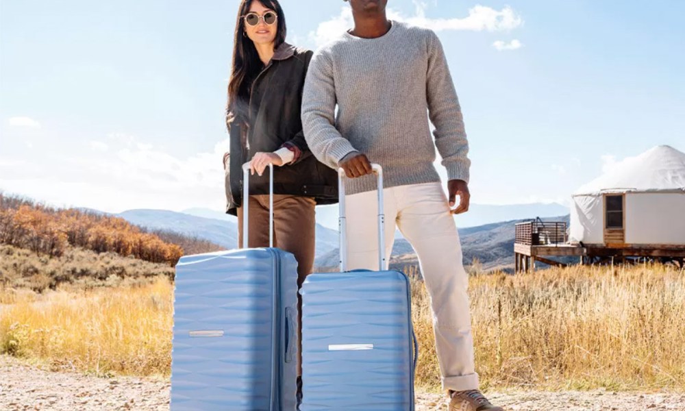 A couple stand in nature with Samsonite suitcases.