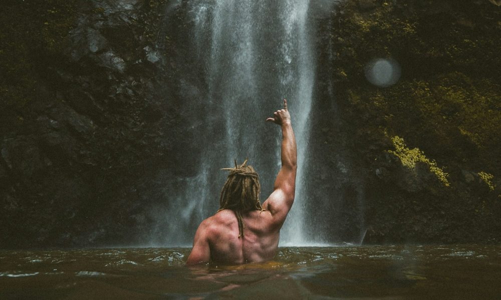 Shirtless man fist in the air punching air by a waterfall outside in nature back muscles