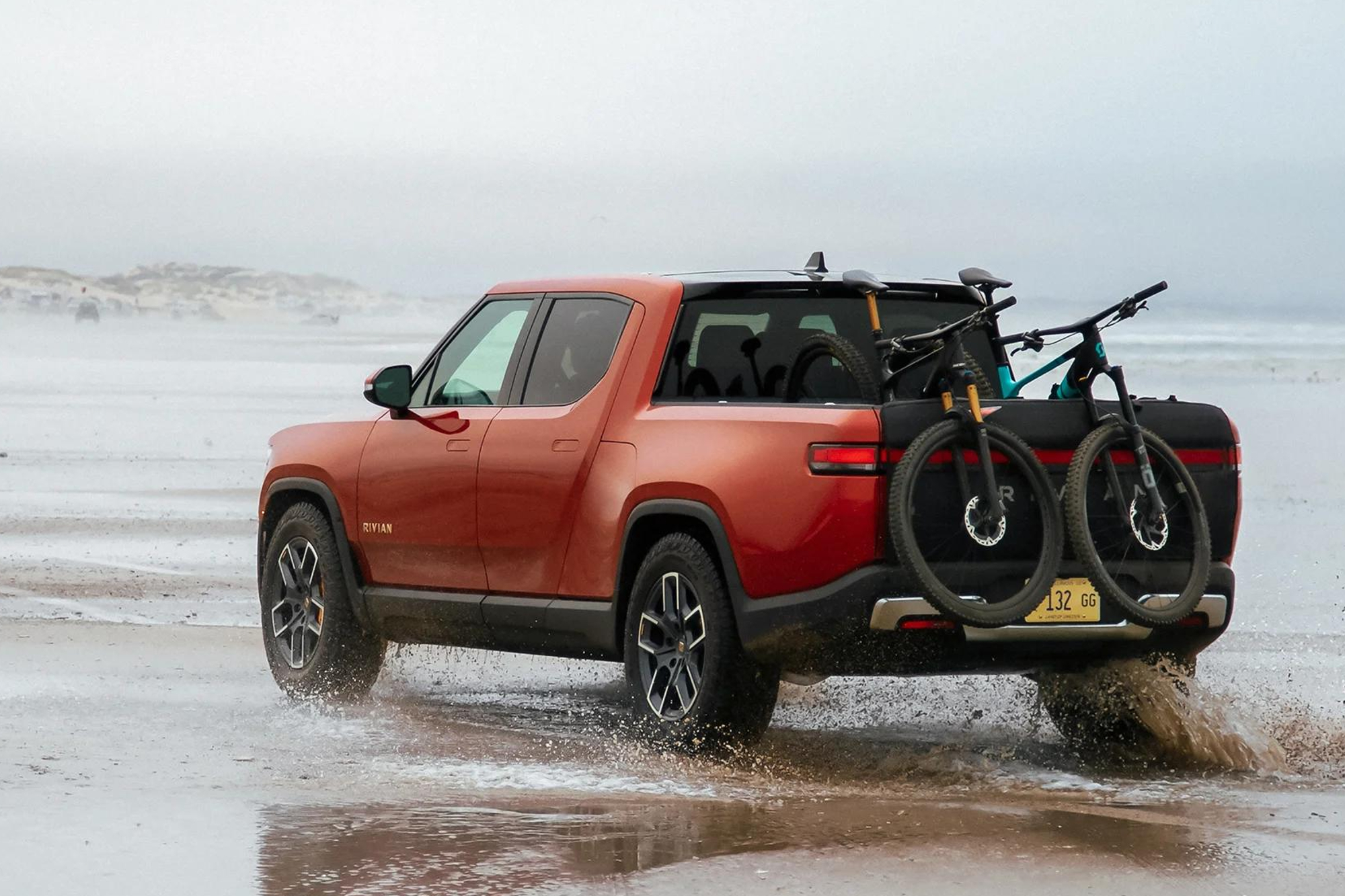 Red second-generation Rivian R1T right rear three-quarter view parked on beach edge facing water bike on back of the truck.