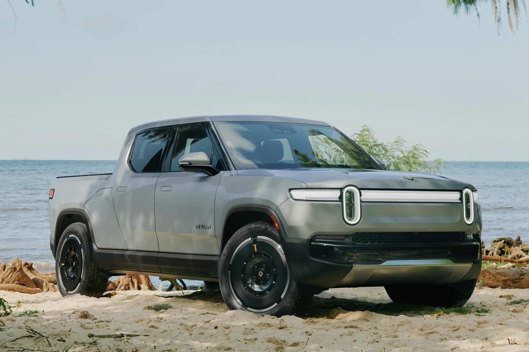 Second-generation Rivian R1T right front three-quarter view parked on a beach backed up to the rocks on the edge of the shore.