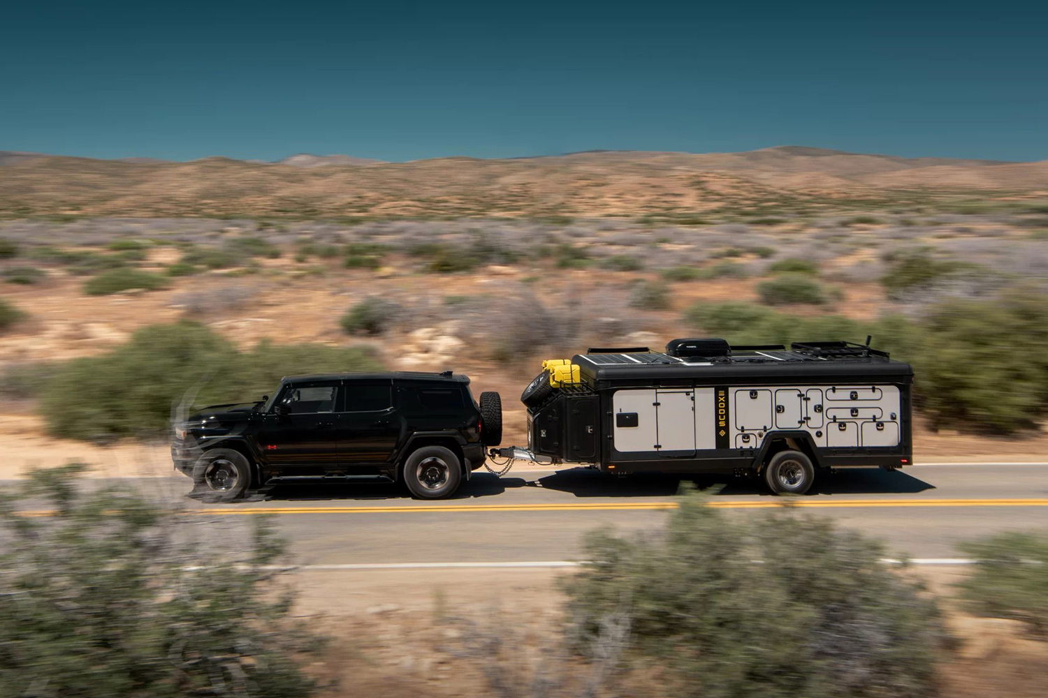 Exodus Capax travel trailer being towed by a black SUV down a desert highway.