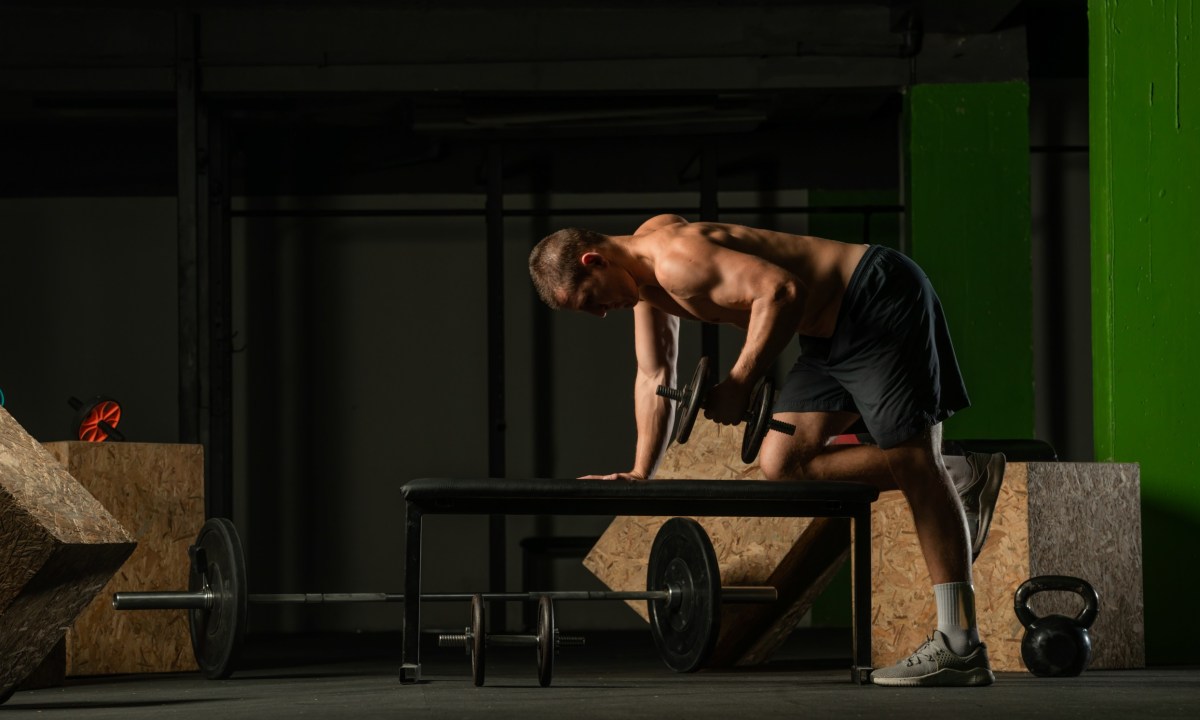 Adobe Shirtless man kneeling on bench in gym doing tricep kickbacks exercise weight dumbbell