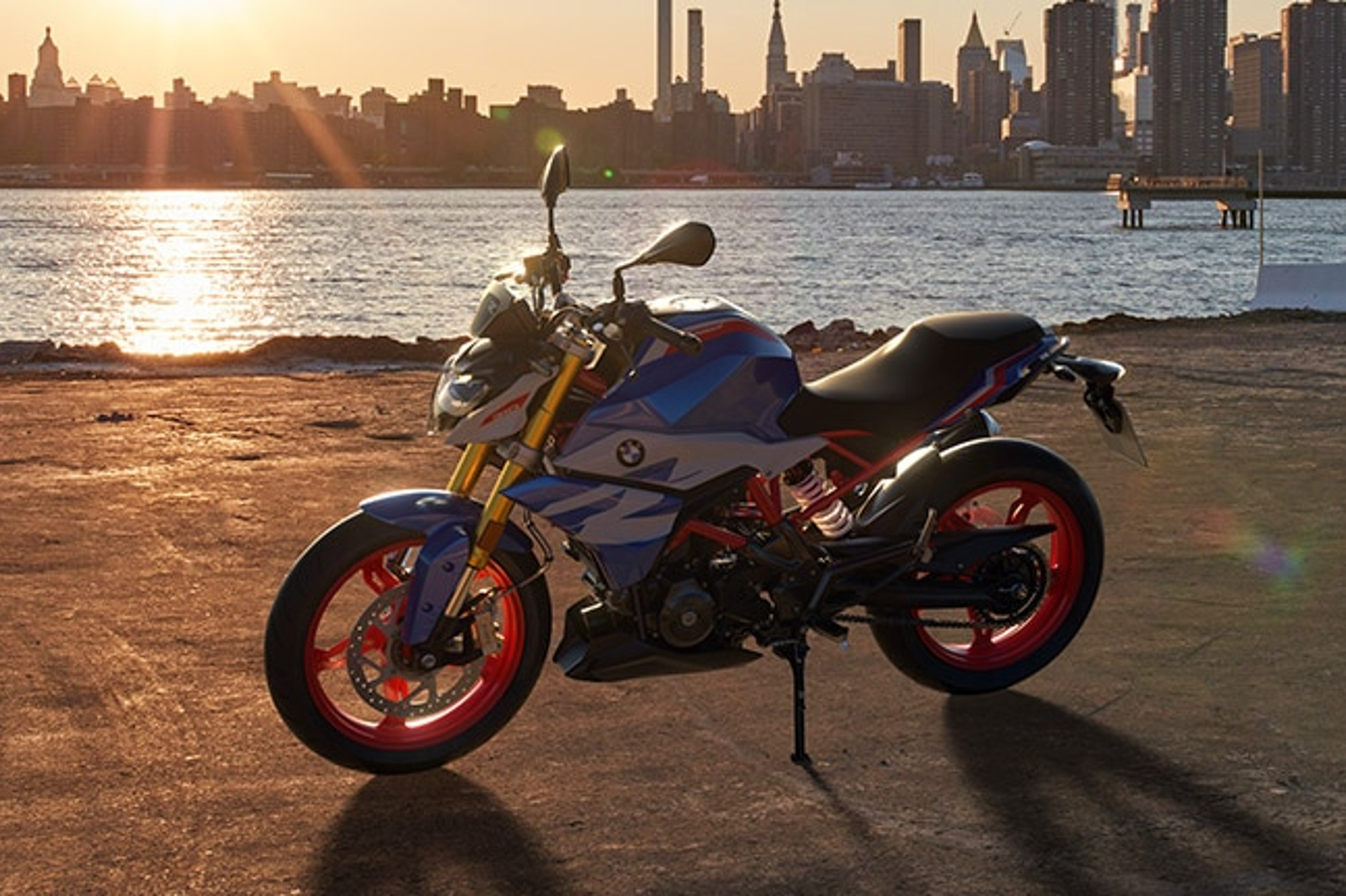 BMW G 310 R parked on pavement with a river and city skyline in the background.