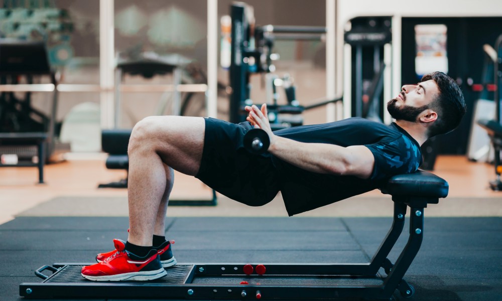 Man wearing black and red sneakers in gym doing hip thrusts with weight barbell on bench