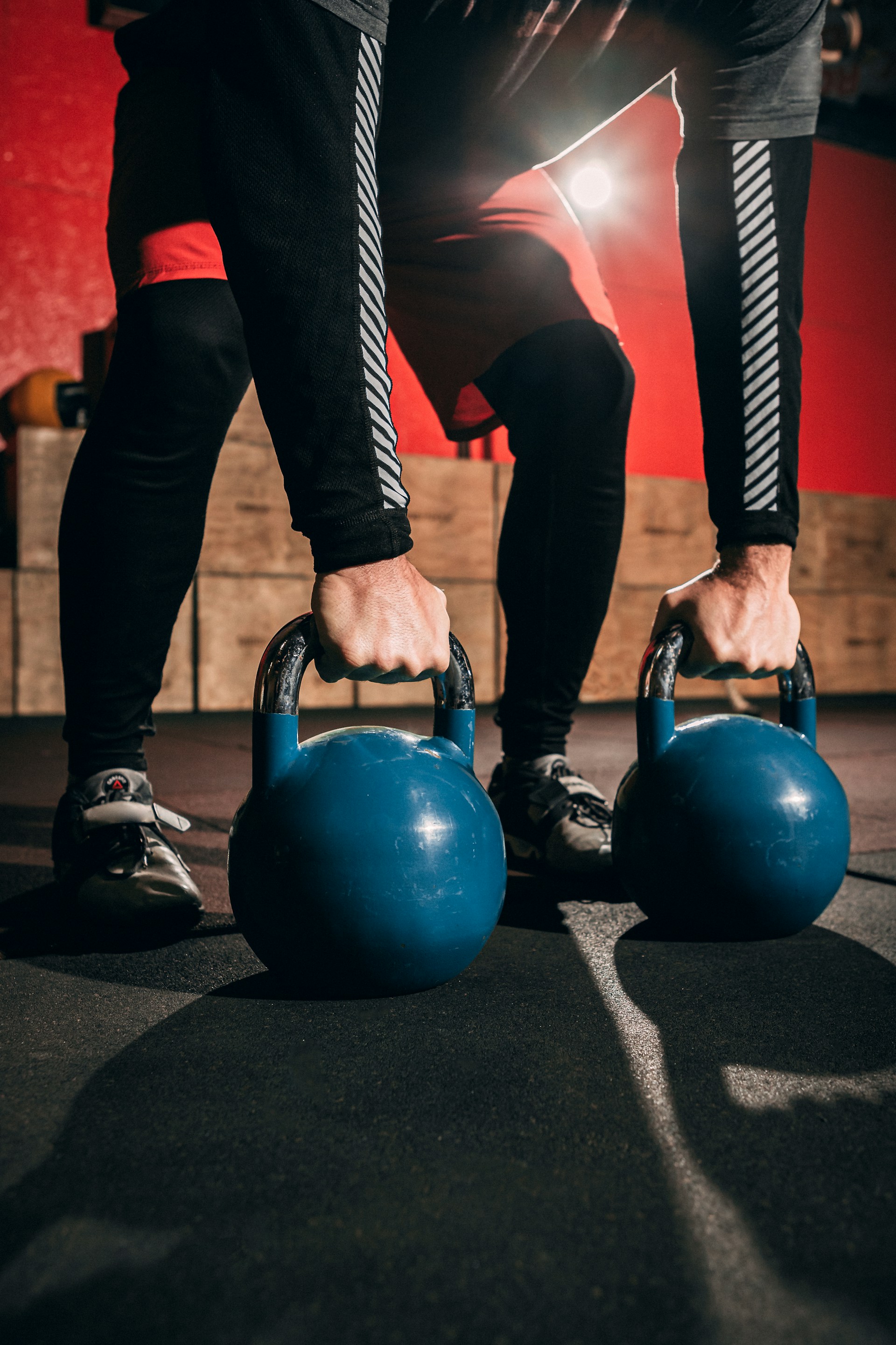 Double kettlebell swing man in gym picking up two blue kettlebells with red background