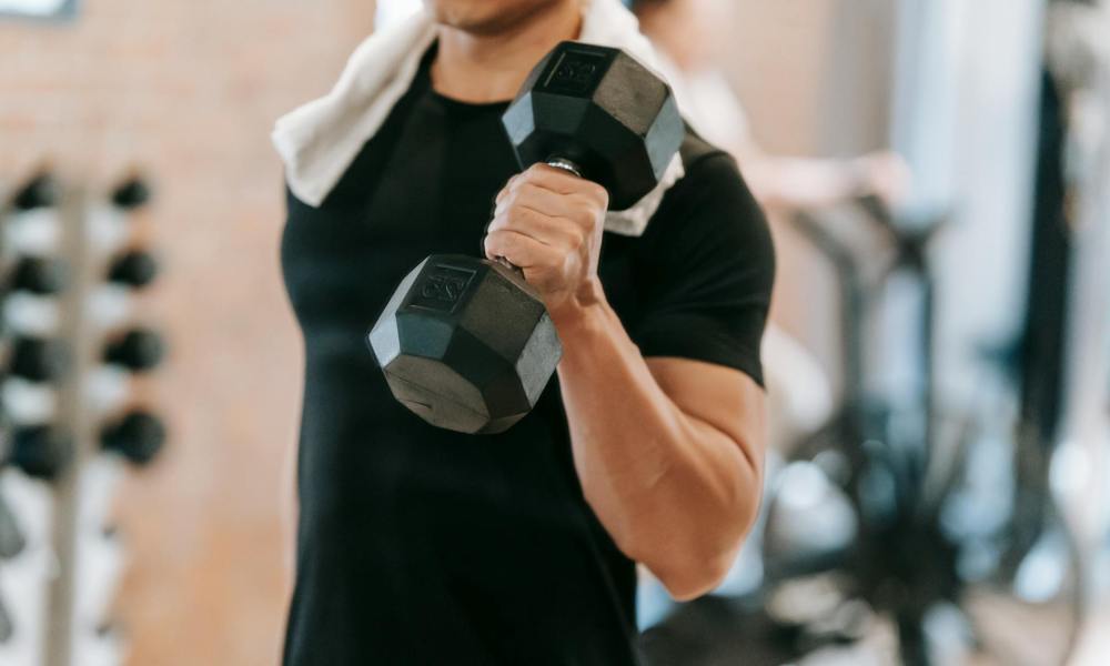 Man wearing a black T-shirt at the gym with a towel around his neck holding a dumbbell doing one arm exercise curl