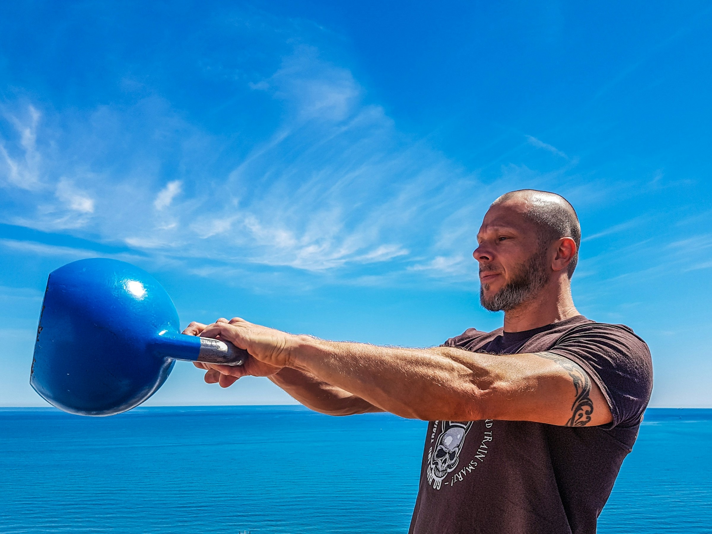 Man swinging blue kettlebell outside by blue ocean and blue sky