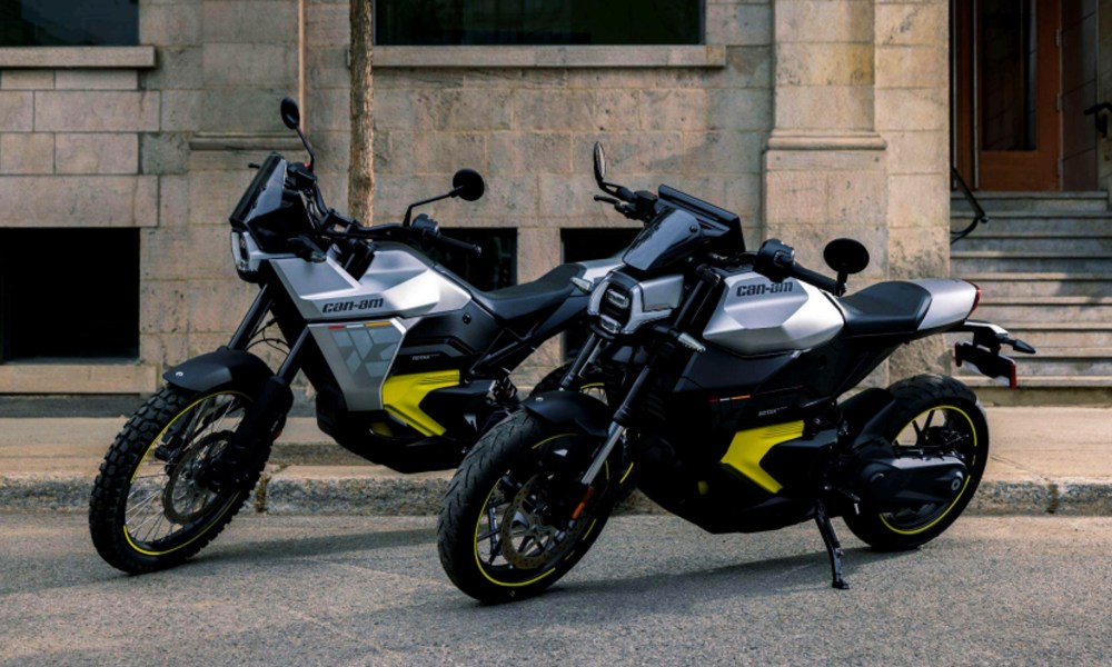 Can-Am Origin and Pulse electric motorcycles parked on pavement in front of a stone building.