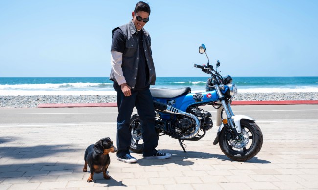 A person with a small dog stnding by a 2025 Honda Dax 125 parked on a beach with the ocean in the background.