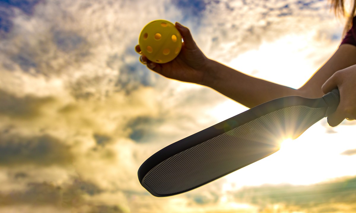 Woman holding a Pickleball paddle and ball as she prepares to serve with the sun rising behind her.