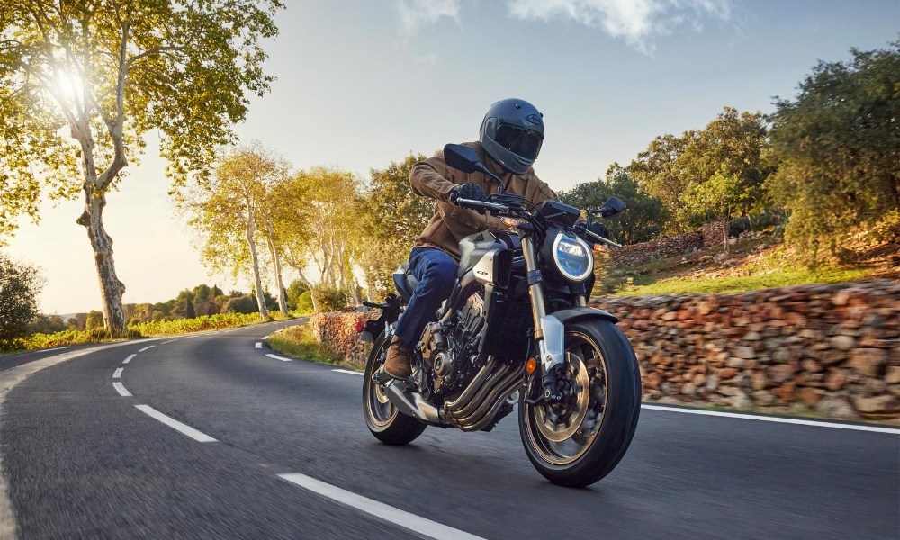 A person riding a Honda CB650R on a countryside road with rock walls, grass, and trees