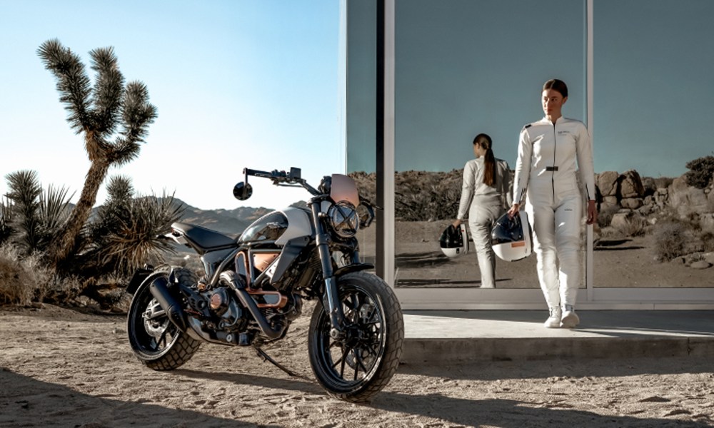 A rider in white looking at a Ducati Scrambler 10th Anniversario Rizoma Edition standing in fromt of a glass-walled desert house.