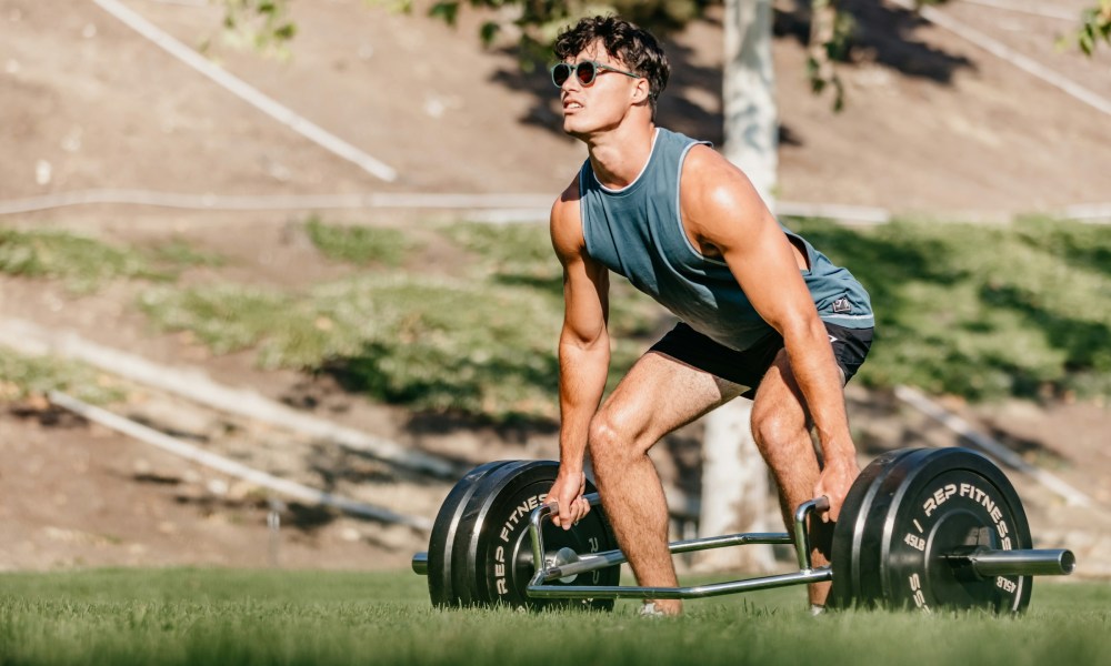 Man outside wearing blue sleeveless shirt doing trap bar deadlift in nature