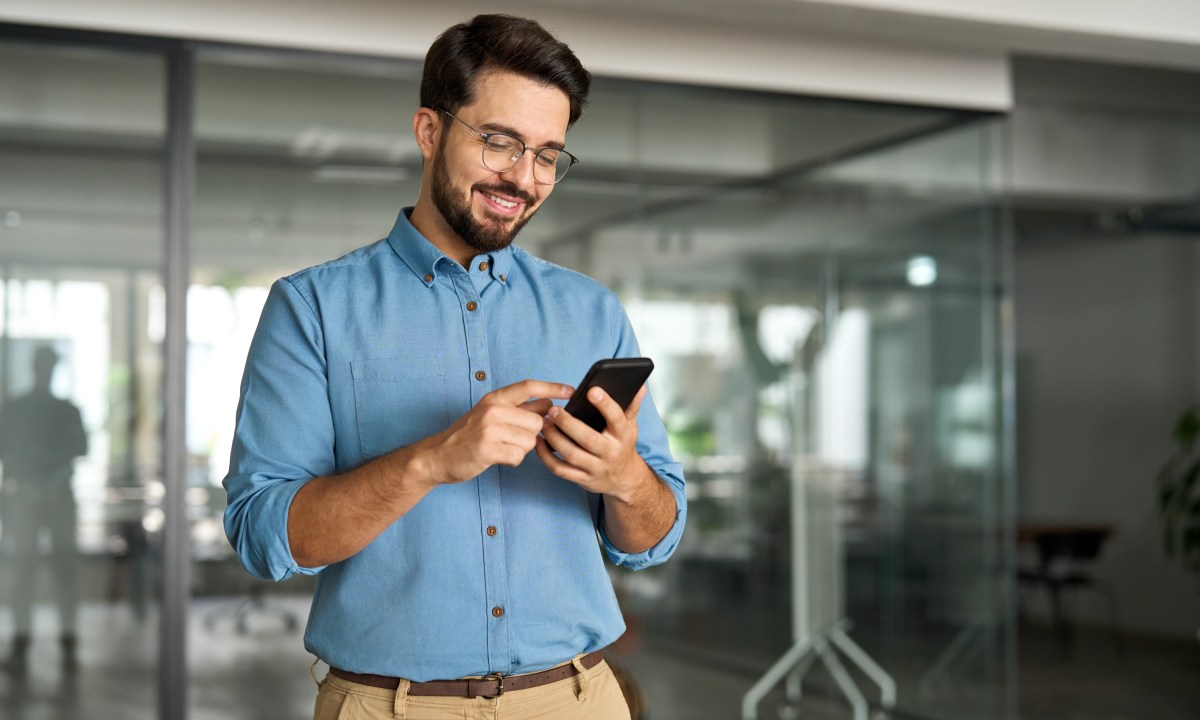 Happy young latin business man holding smartphone standing in office