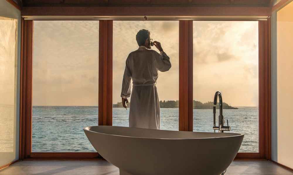 man in a white robe in hotel looking out window to the ocean