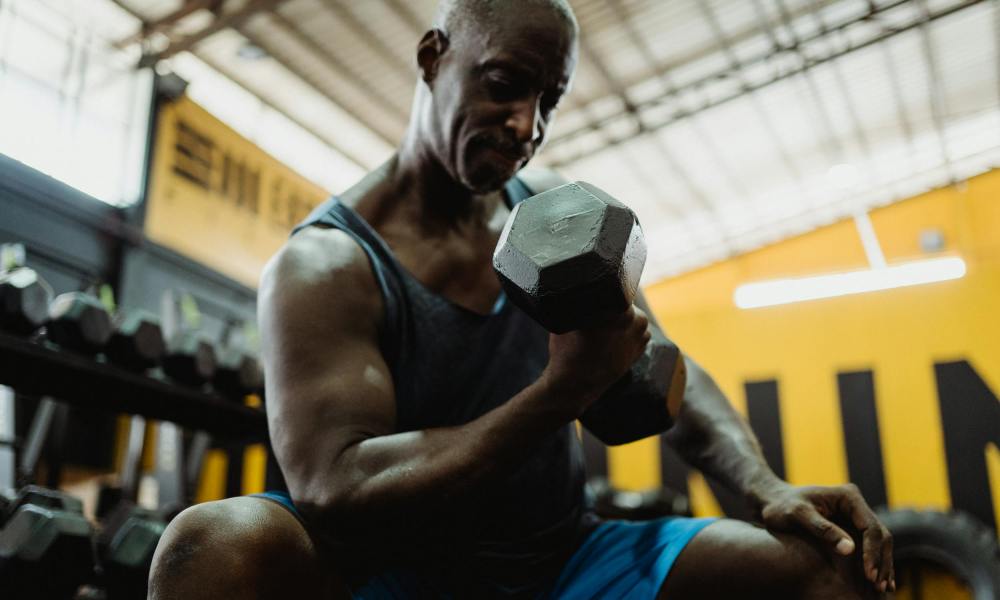 Man wearing blue tank sitting in gym doing dumbbell concentration curl on bench