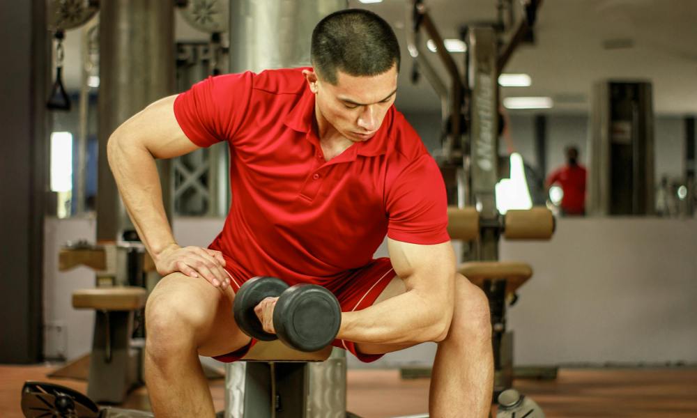 man in gym wearing red shirt doing concentration curl with dumbbell seated on bench