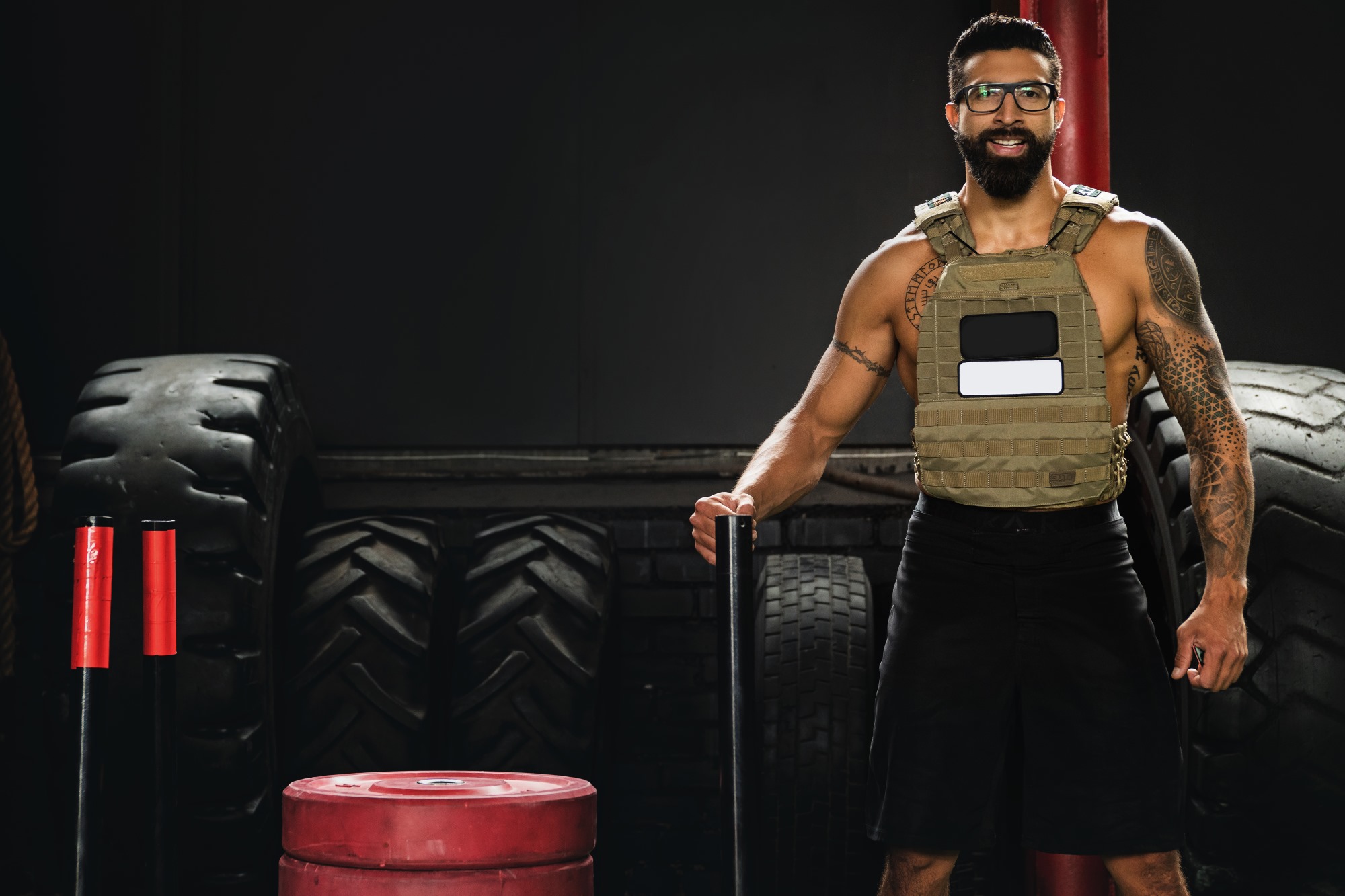 man wearing weighted vest and glasses smiling in gym next to tires