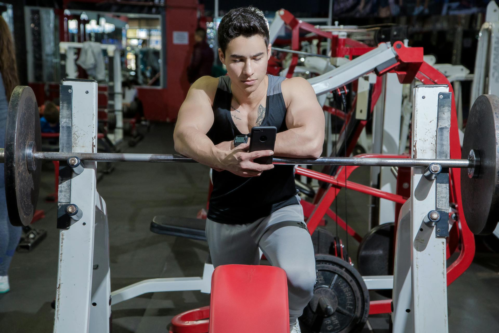 man at gym on phone hogging machine leaning on barbell