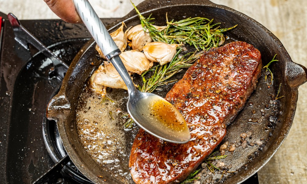 A person is cooking a sirloin steak in a pan with herbs and spices. The steak is brown and he is well-cooked